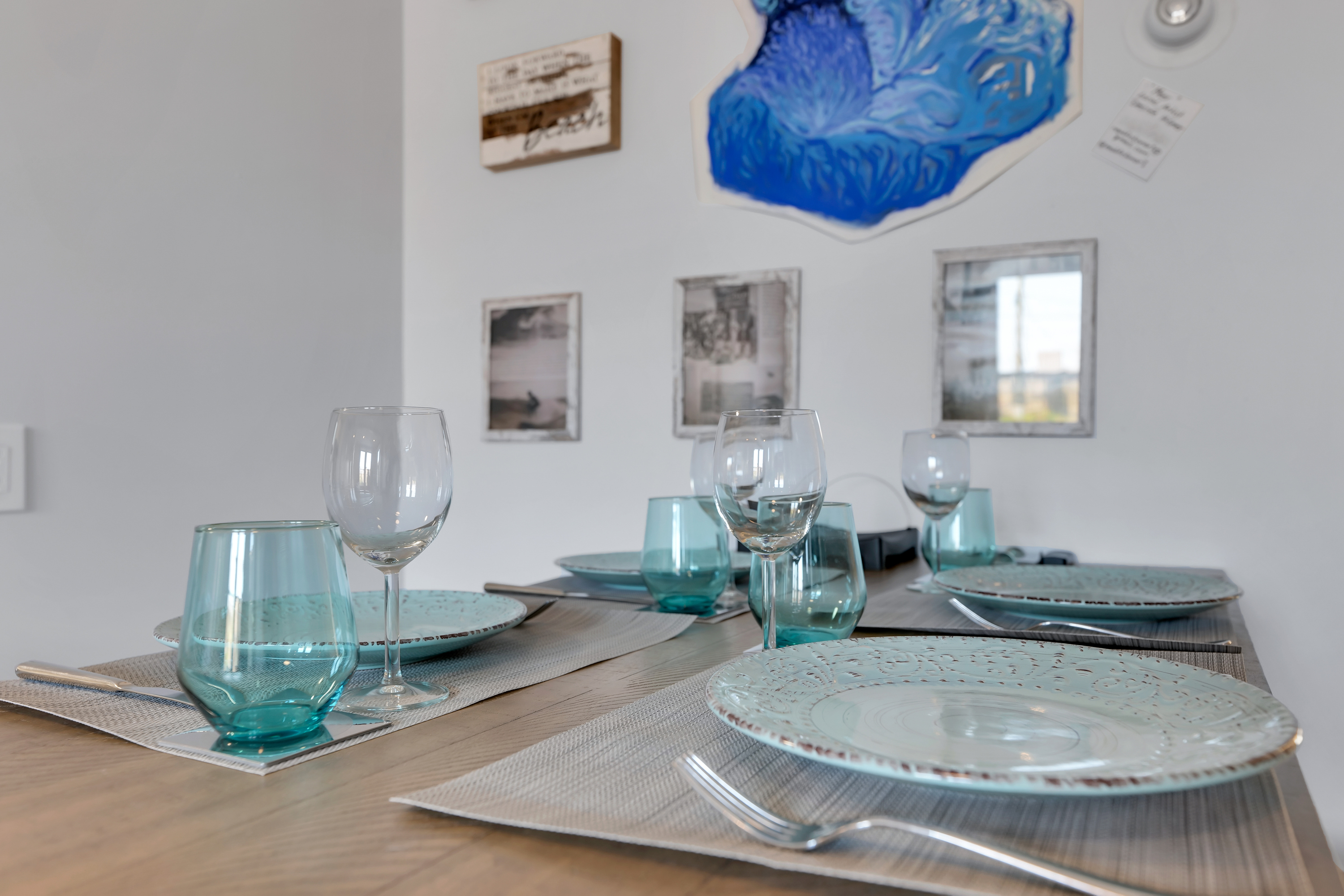 A close-up of a dining table set for four with blue-tinted glasses, wine glasses, patterned plates, and neutral placemats—perfect for Charleston rental marketing. Art and photographs decorate the white wall in the background.