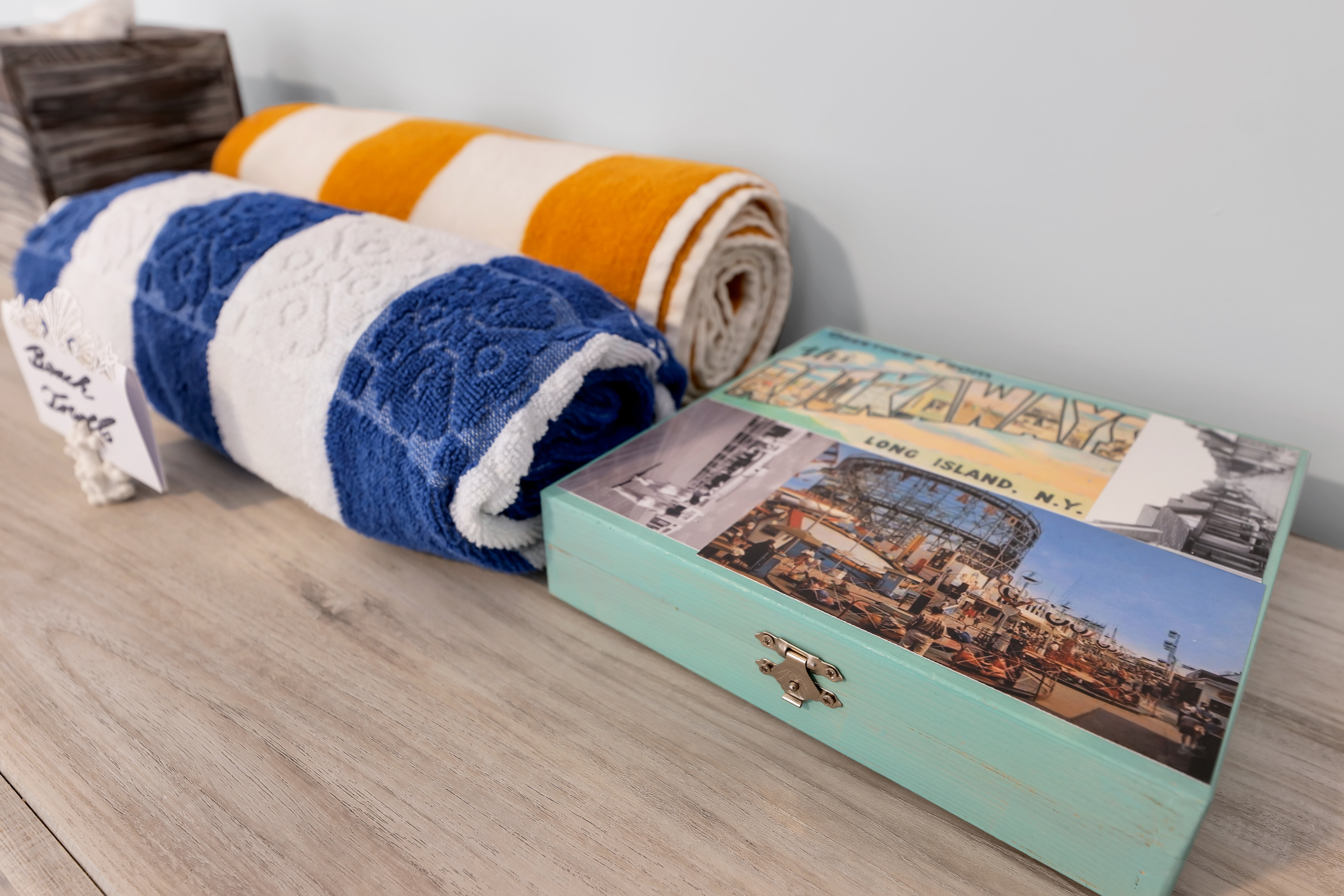 Two rolled beach towels, one blue and white and one yellow and white, sit on a wooden surface next to a decorative box with vintage Long Island, NY imagery—perfect for capturing that inviting Charleston rental marketing aesthetic.