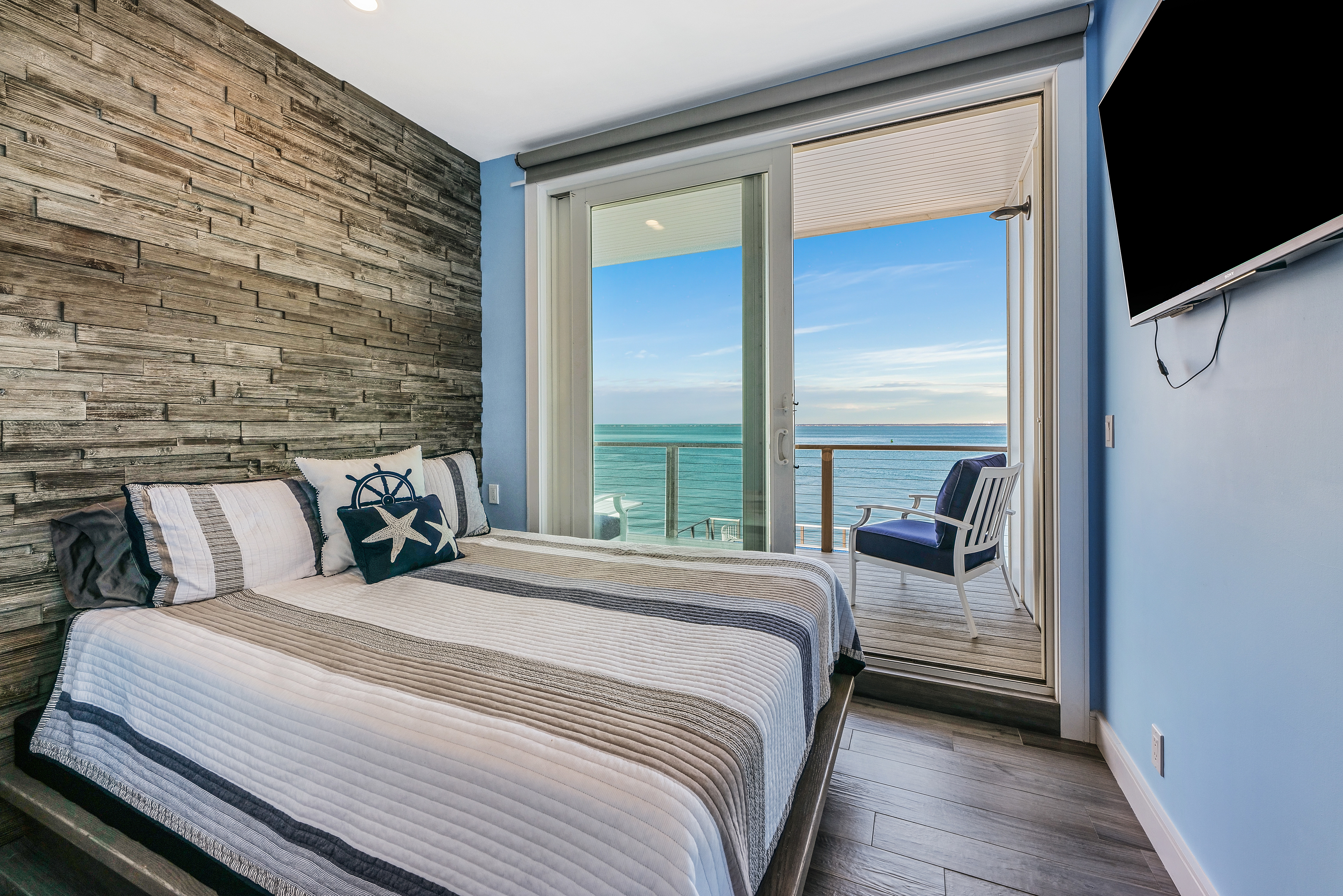 Modern bedroom with a striped bedspread, nautical-themed pillows, and rustic wood accent wall. Perfect for any Staging Guide, glass doors open to a balcony with a chair, offering scenic ocean views under blue skies. TV mounted on the wall.