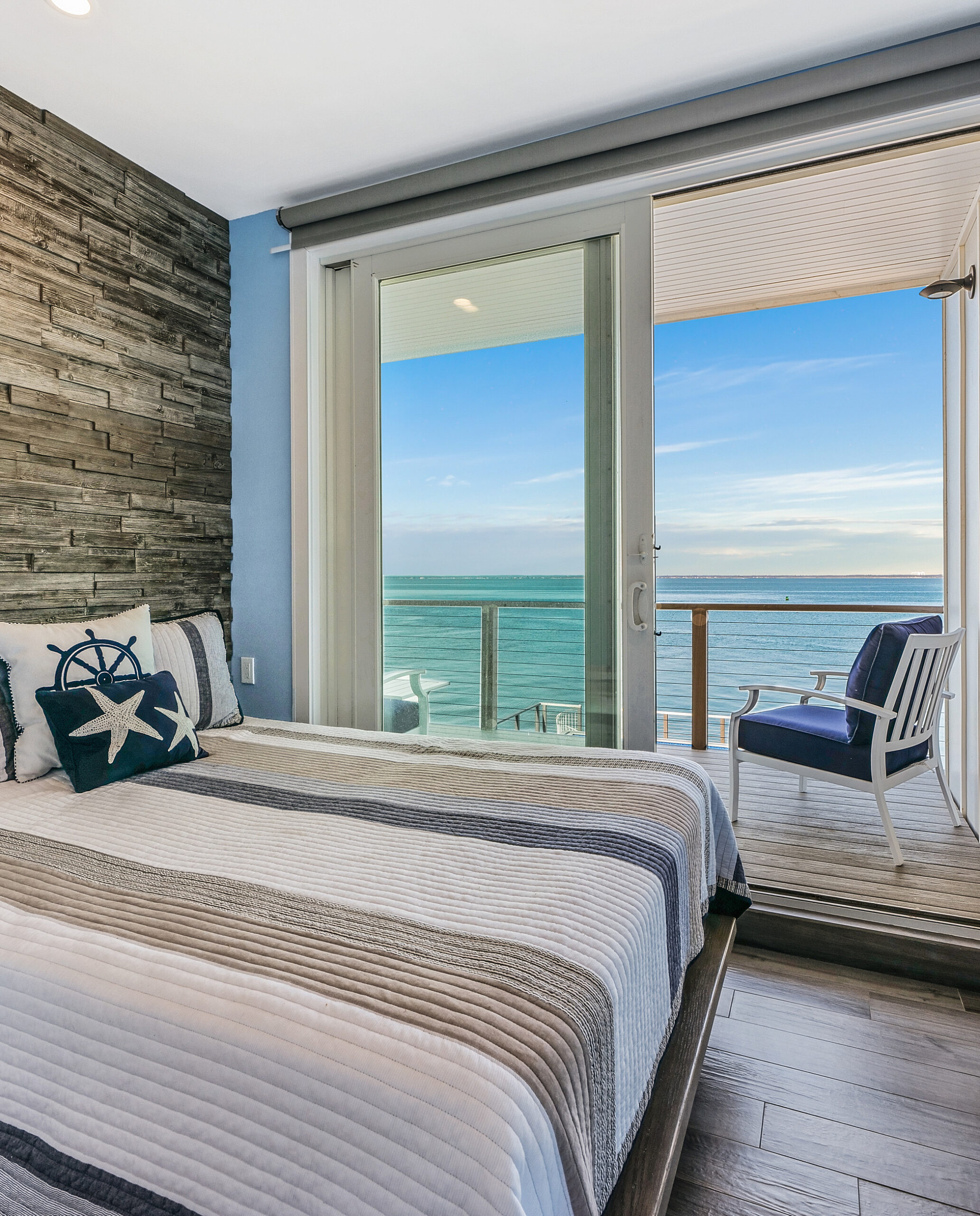 A cozy bedroom with a nautical theme features a bed with striped bedding and a starfish pillow. According to any good staging guide, glass doors open to a balcony with chairs and a scenic ocean view under a blue sky.