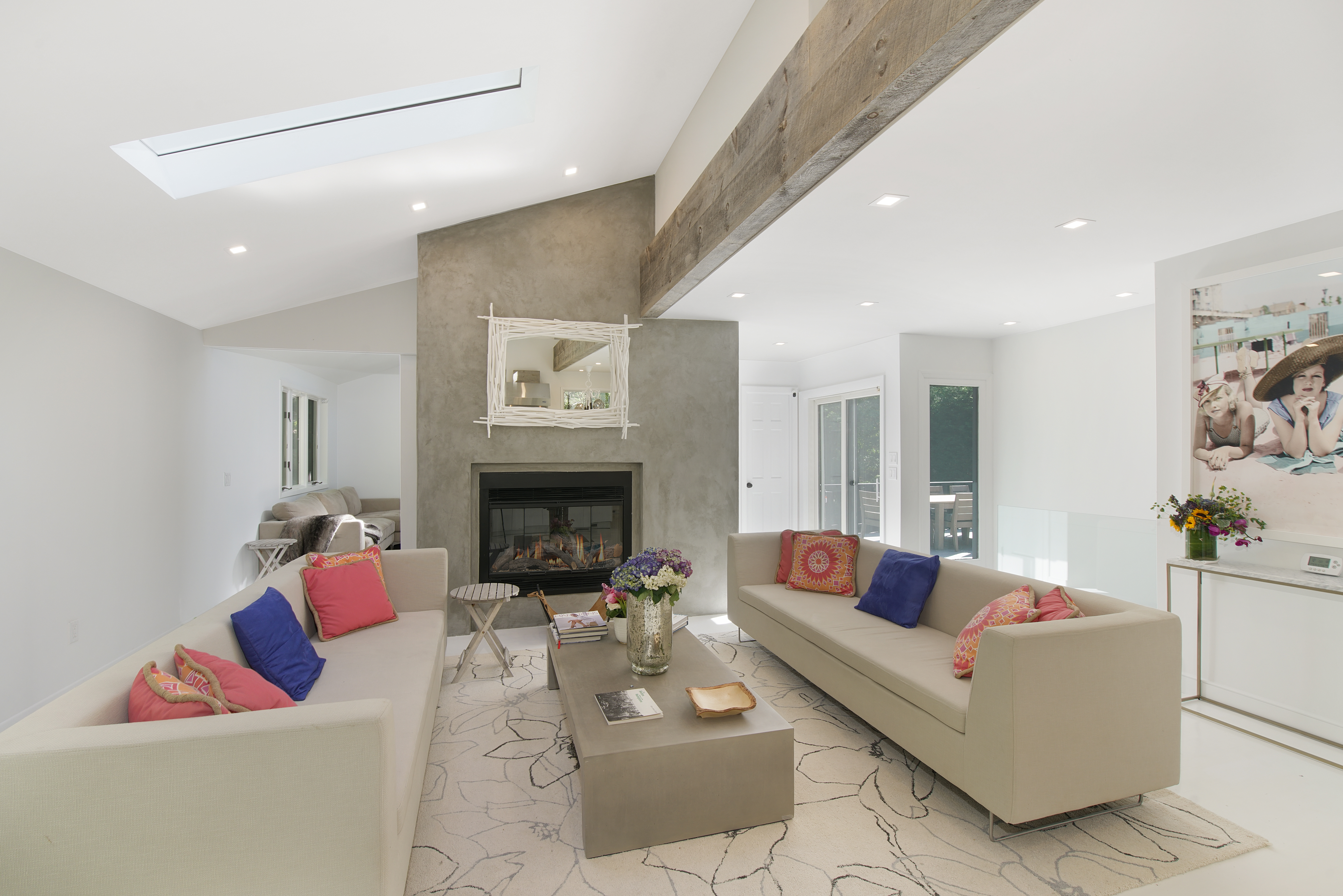 Modern living room perfect for Charleston rental marketing, featuring light walls, two beige sofas with colorful cushions, a central coffee table, a fireplace with a mirror above it, exposed wooden beam, and striking large wall art in the background.