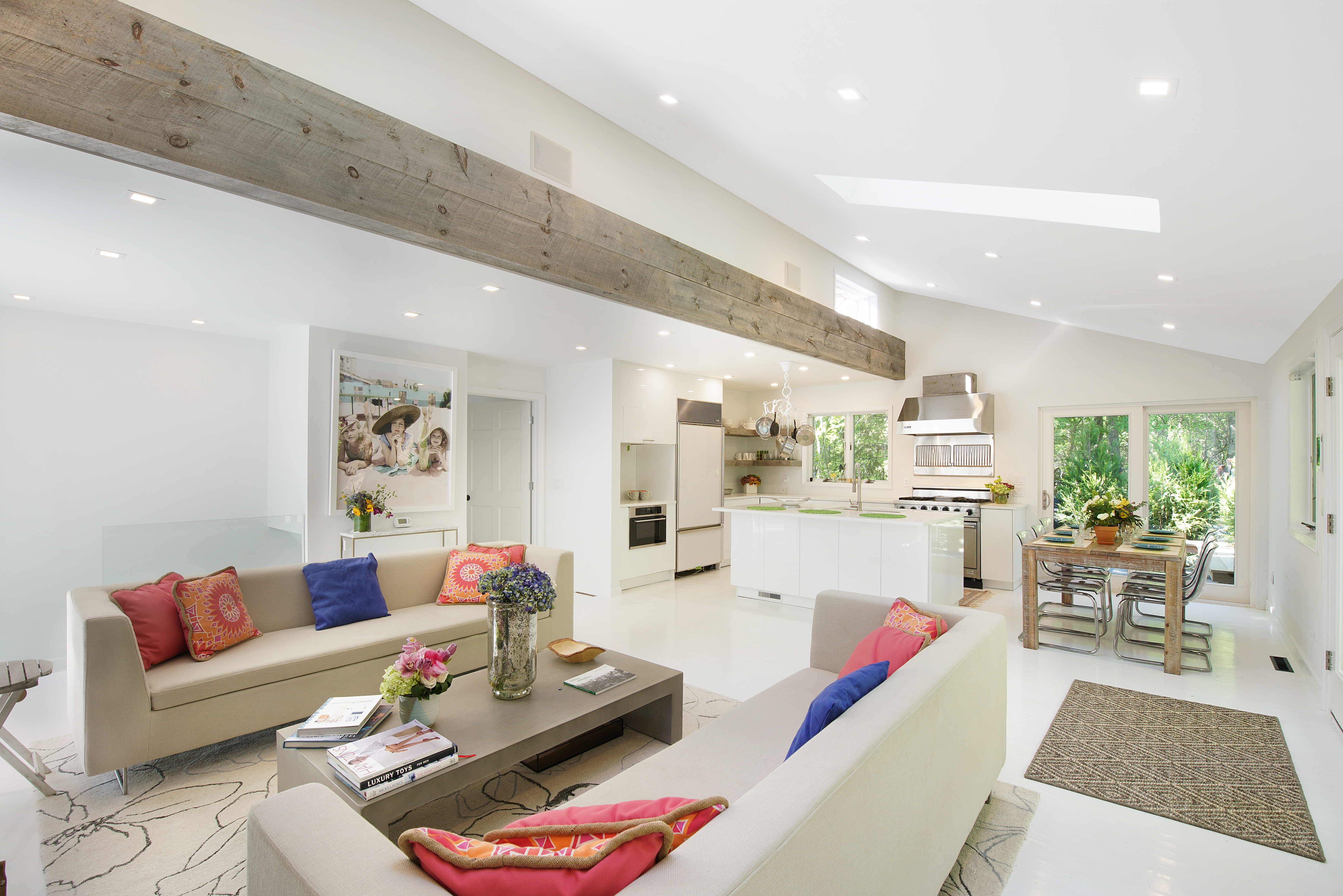 A bright, modern open-plan living space perfect for Charleston rental marketing, featuring two beige sofas, colorful cushions, a coffee table, and a kitchen and dining area in the background. Large windows and skylights flood the room with natural light.