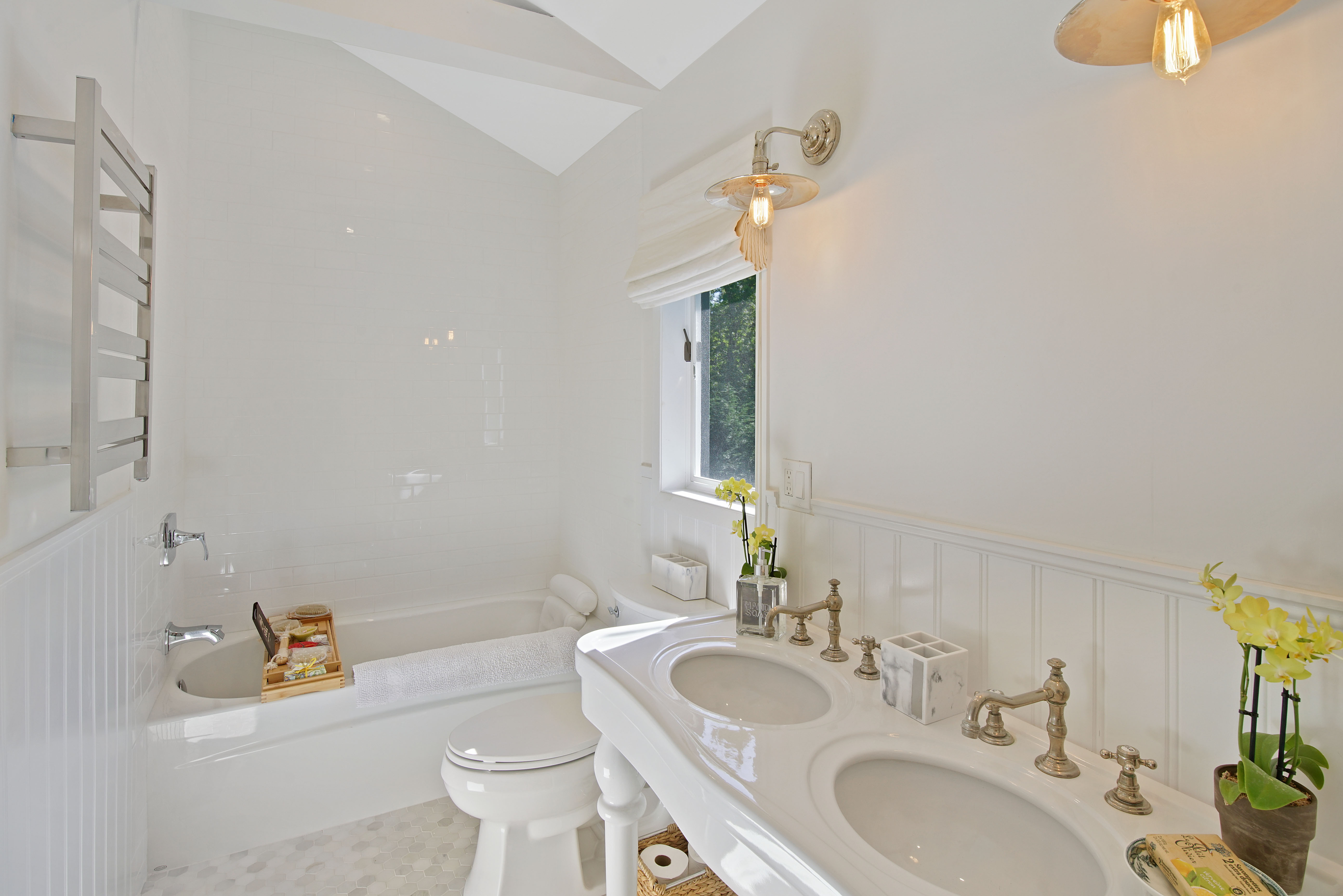 Bright, white bathroom ideal for Charleston rental marketing, featuring a double sink vanity, brass fixtures, a towel rack, a bathtub, a window with Roman shade, decorative yellow flowers, and wall-mounted light fixtures.