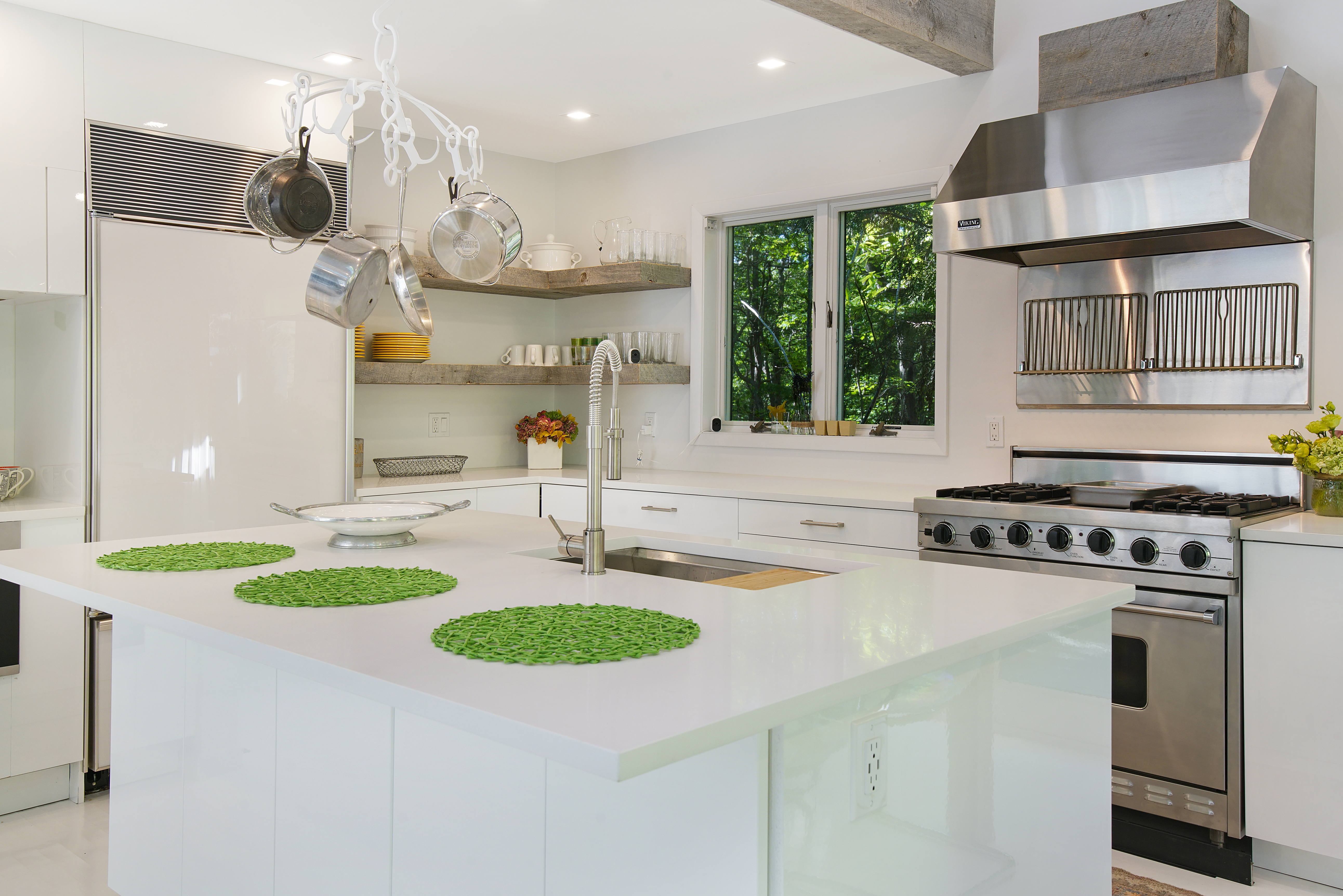 Modern white kitchen with stainless steel appliances, a large island featuring a sink, and green placemats—perfect for Charleston rental marketing. Pots hang above the island, with open shelves and dishes near a sunlit window.