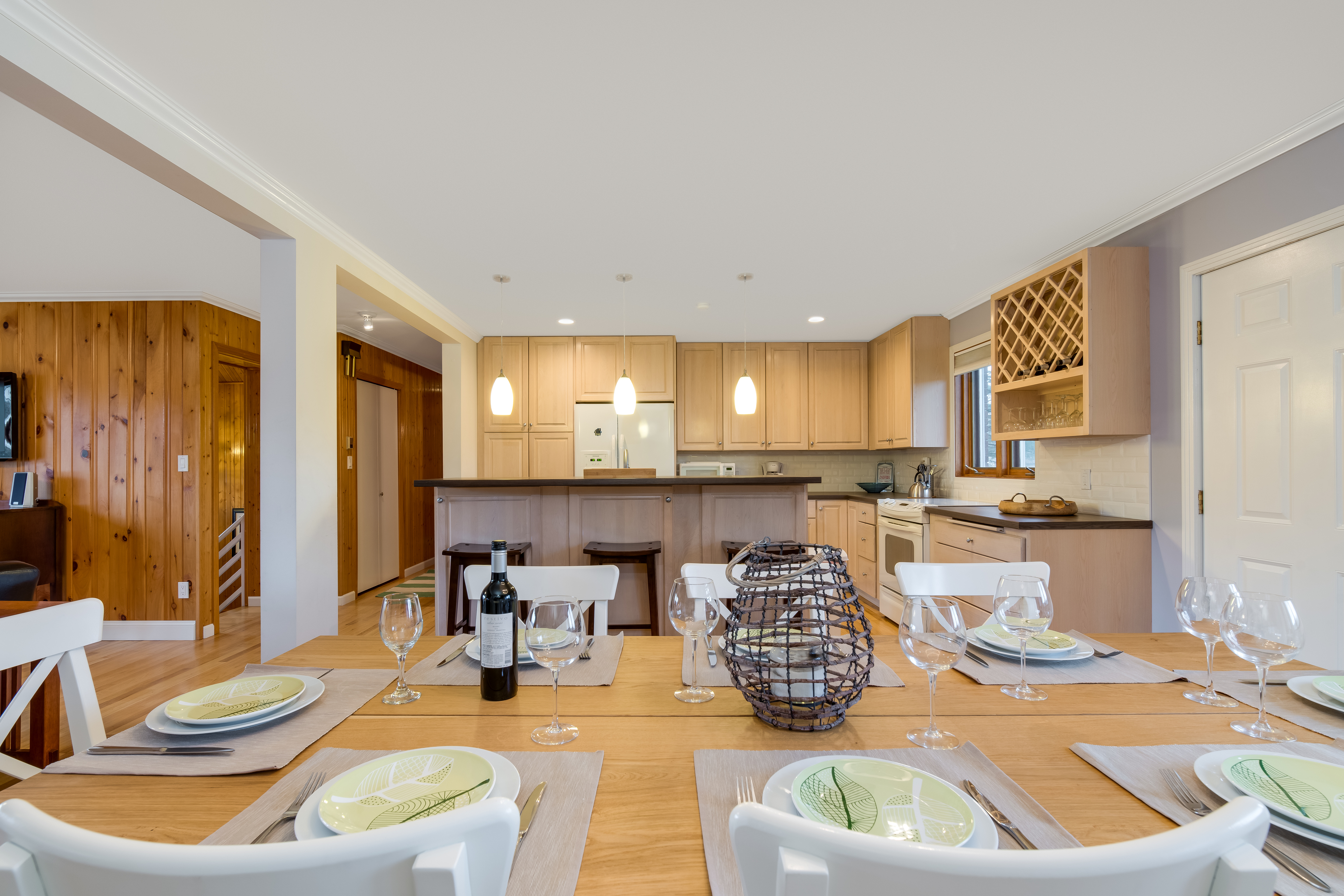 A modern open-concept kitchen and dining area, perfect for Charleston rental marketing, features light wood cabinets, a breakfast bar with stools, a set dining table, wine glasses, a bottle of wine, and a decorative centerpiece.