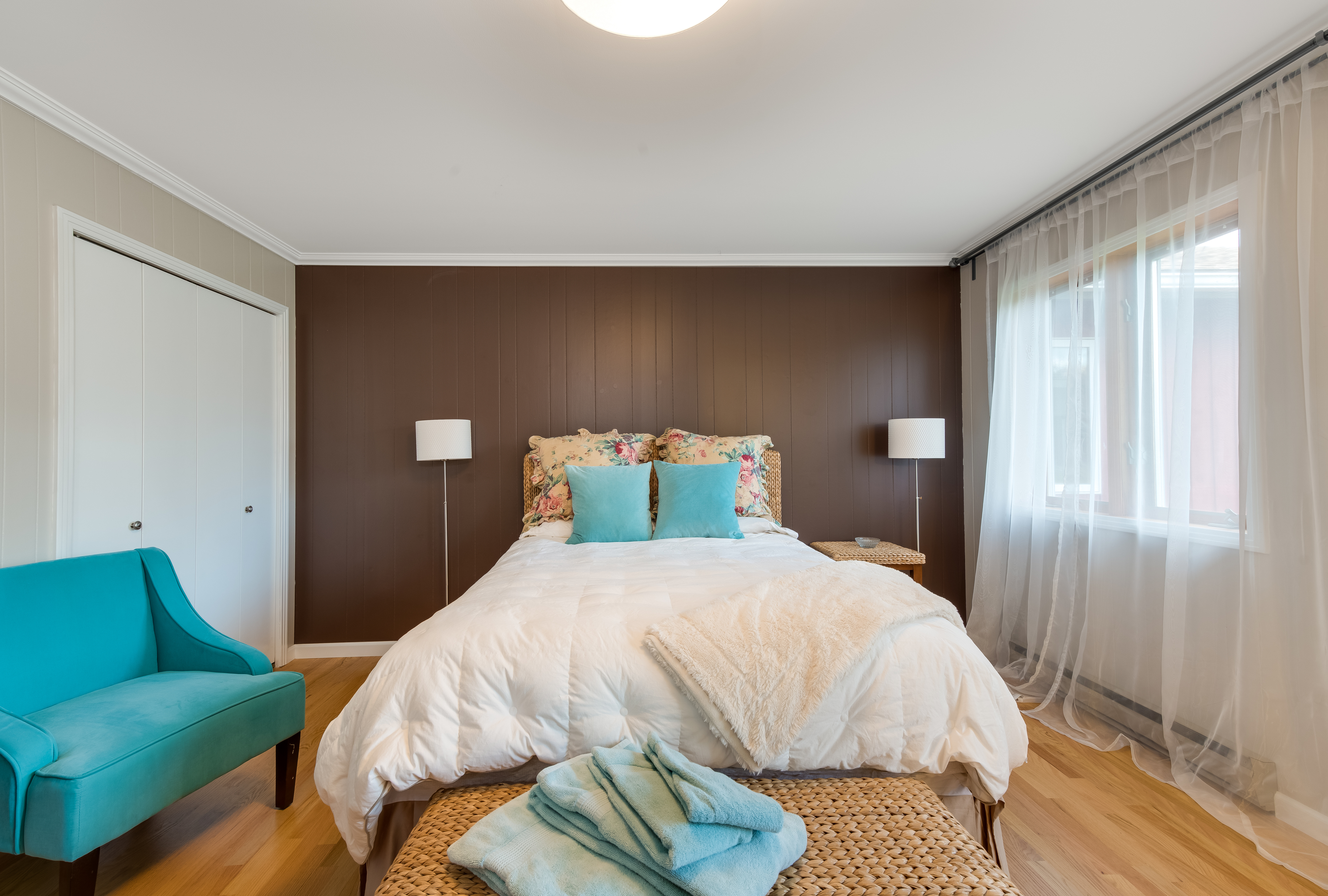 A cozy bedroom perfect for Charleston rental marketing, featuring a double bed with floral and teal accent pillows, a teal armchair, two bedside lamps, a wicker bench with towels, and sheer curtains on a large sunlit window.