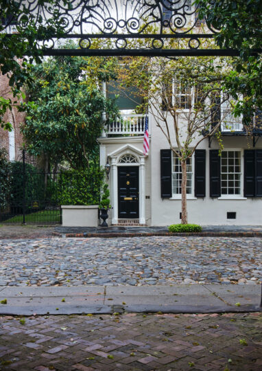 View through an ornate wrought iron gate, looking across a cobblestone street toward a historic white house—perfect imagery for Charleston rental marketing—with black shutters, a black door, American flag, and trees in front.