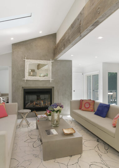 Modern living room perfect for Charleston rental marketing, featuring light walls, two beige sofas with colorful cushions, a central coffee table, a fireplace with a mirror above it, exposed wooden beam, and striking large wall art in the background.
