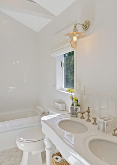 Bright, white bathroom ideal for Charleston rental marketing, featuring a double sink vanity, brass fixtures, a towel rack, a bathtub, a window with Roman shade, decorative yellow flowers, and wall-mounted light fixtures.